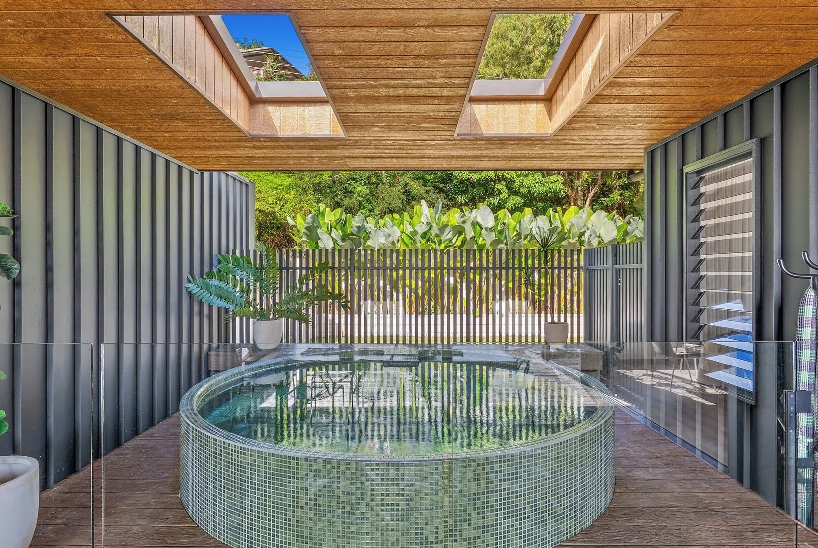 A modern outdoor hot tub area with a wooden ceiling, a glass enclosure, and a metal fence.