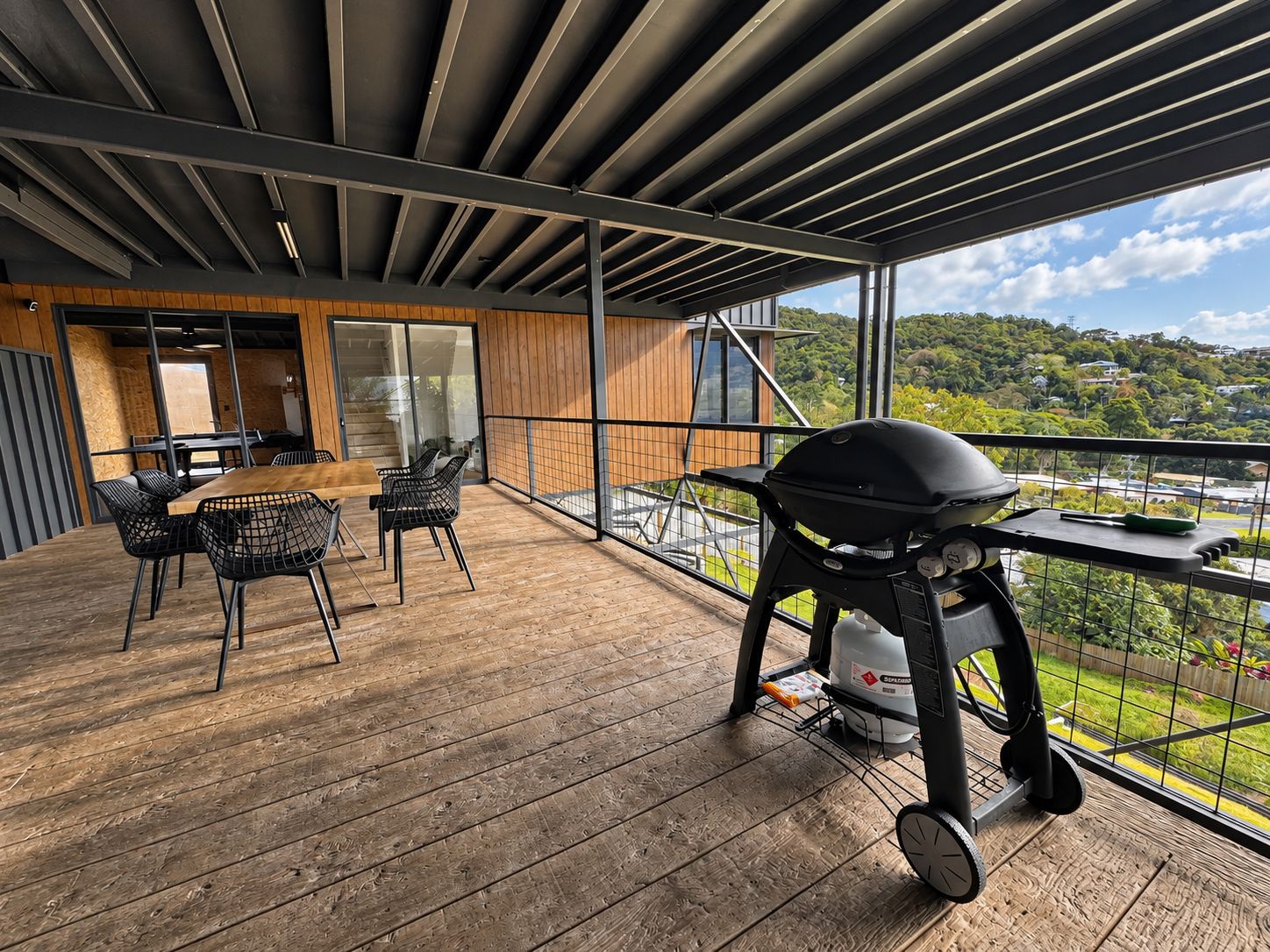 A covered outdoor area with a wooden deck, a barbecue grill, and a dining table with chairs.