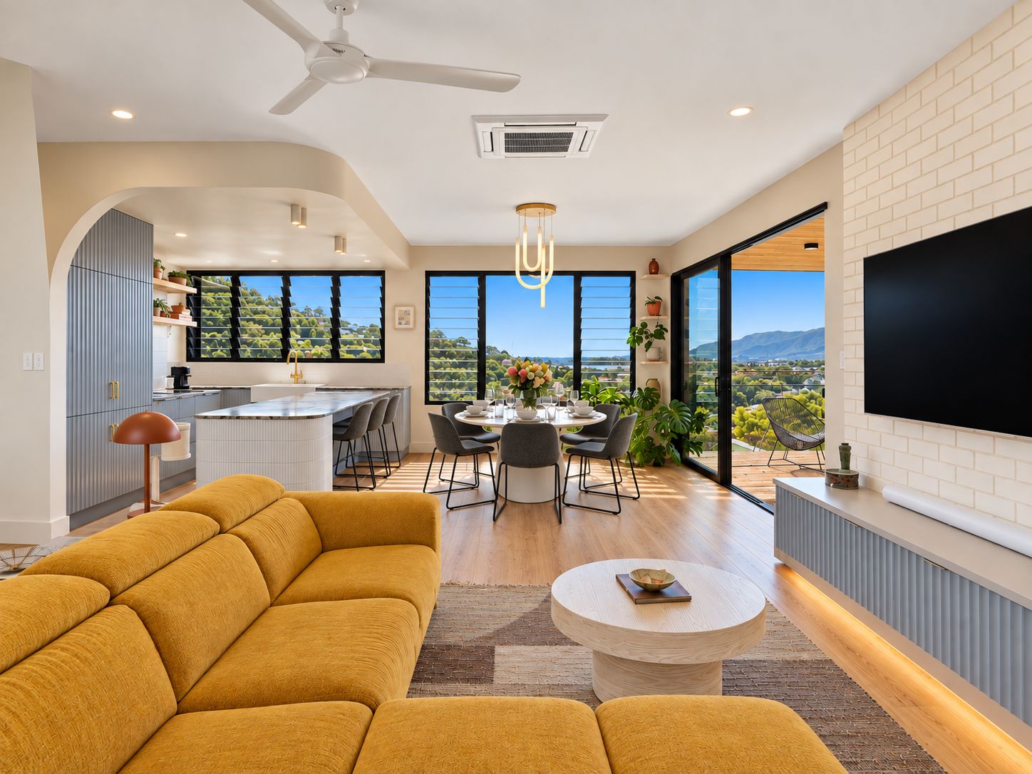 A modern living room with a sofa, a dining area with a table and chairs, a kitchen with a bar, and a large TV on the wall.