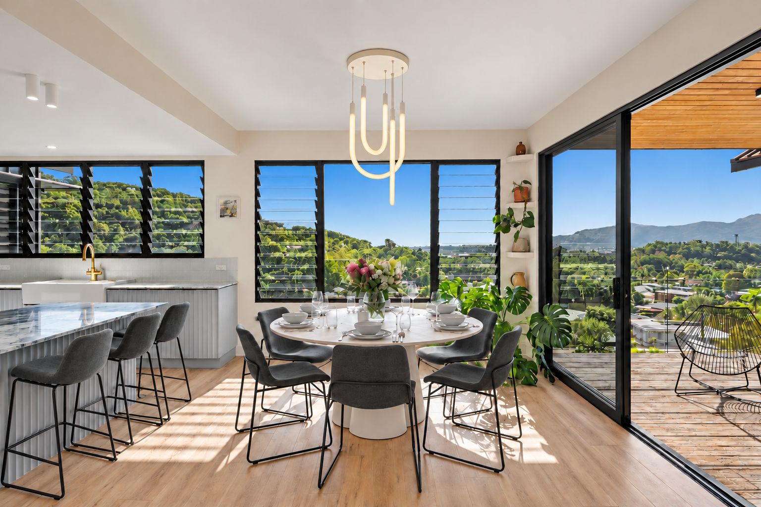 A modern kitchen with a central island, dining area with round table and chairs, and large windows offering a view of greenery and mountains.