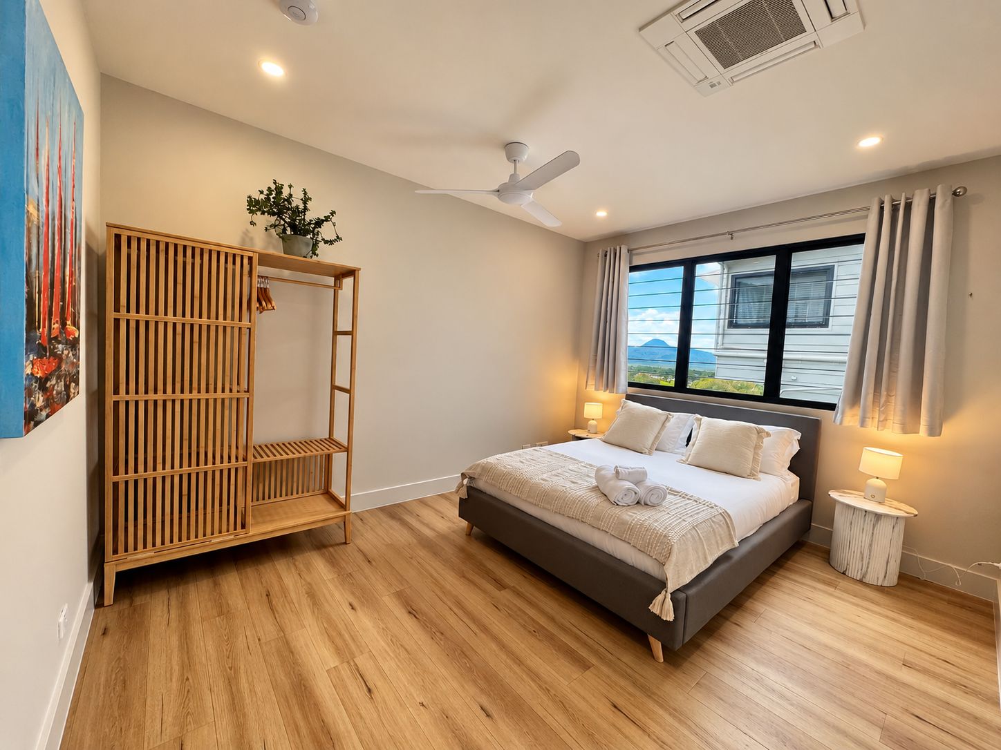 A bedroom with a bed, a wooden wardrobe, a ceiling fan, a window with a view, and a bedside table with a lamp.