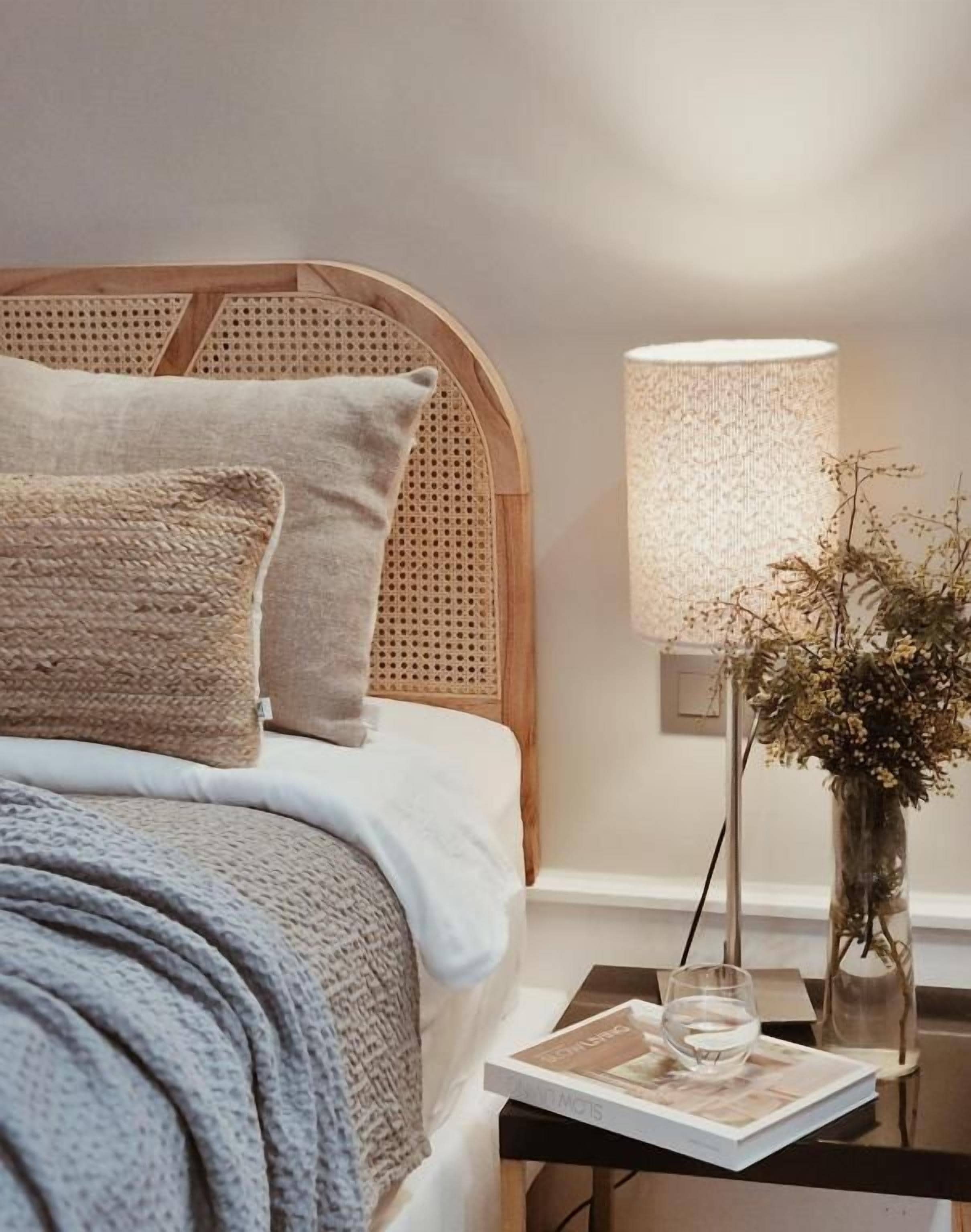 A neatly made bed with a woven headboard, a bedside table with a lamp and a vase of dried flowers, and a book on the table.