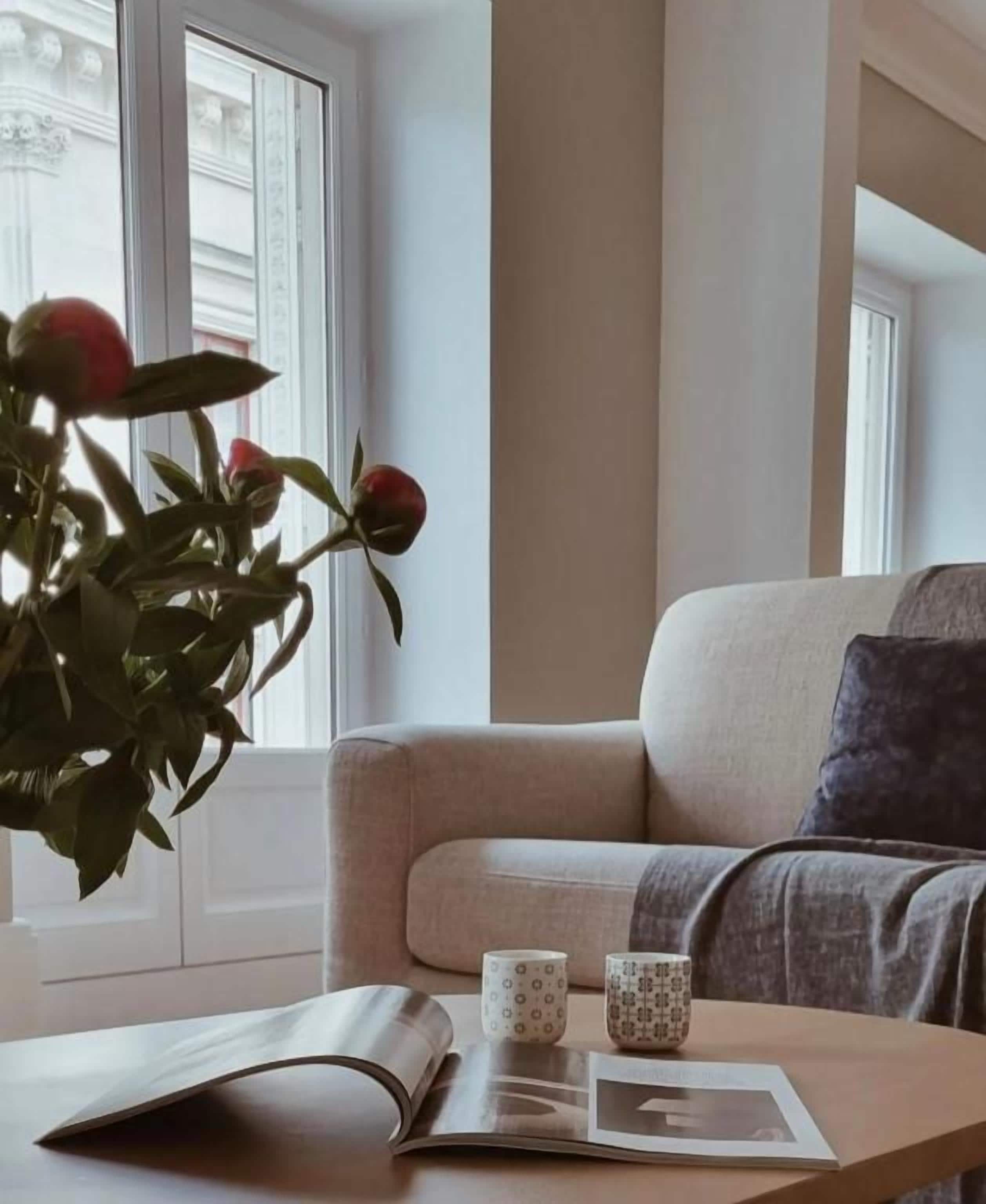 Una habitación con un sofá, una mesa con un libro y dos tazas, y una ventana con una planta.