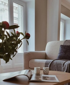 Una habitación con un sofá, una mesa con un libro y dos tazas, y una ventana con una planta.