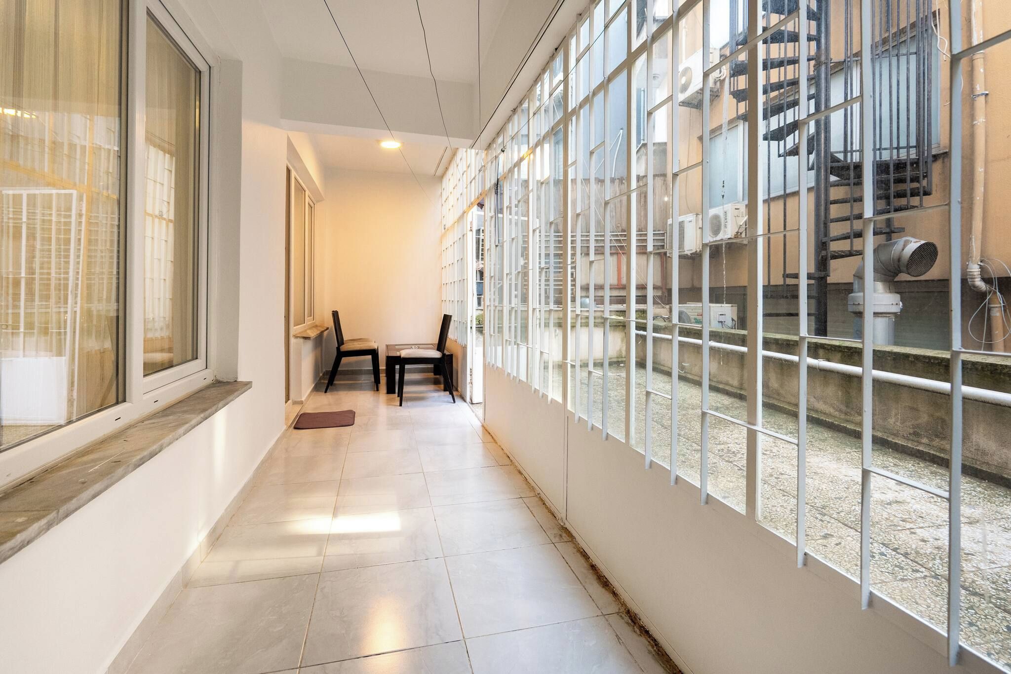 A corridor with a tiled floor, a chair, a table, and a window with a view of an outdoor area with stairs and pipes.
