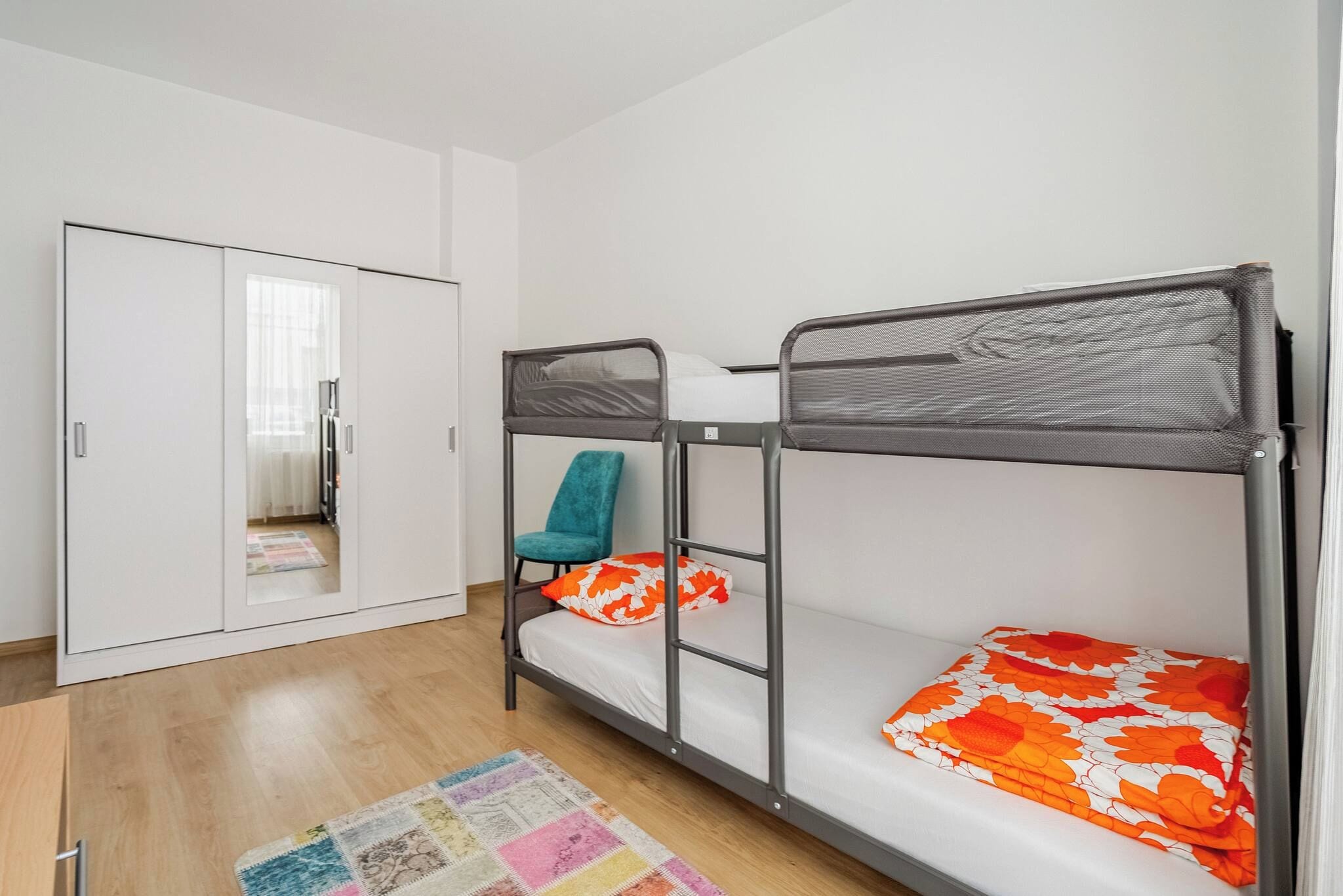A room with bunk beds, a wardrobe, a teal chair, and a colorful rug.
