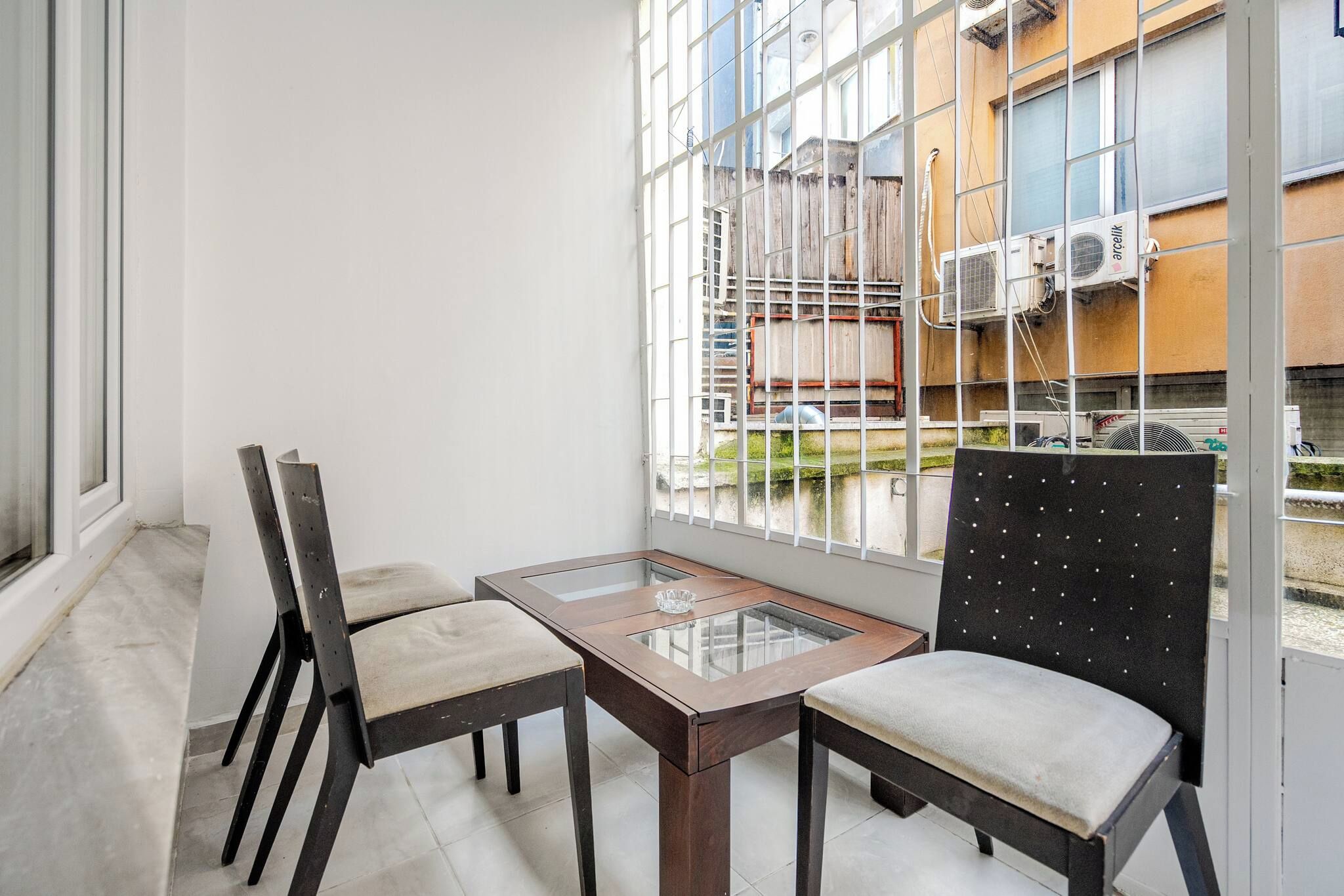 A balcony with a glass table and two chairs, overlooking a building with air conditioning units.