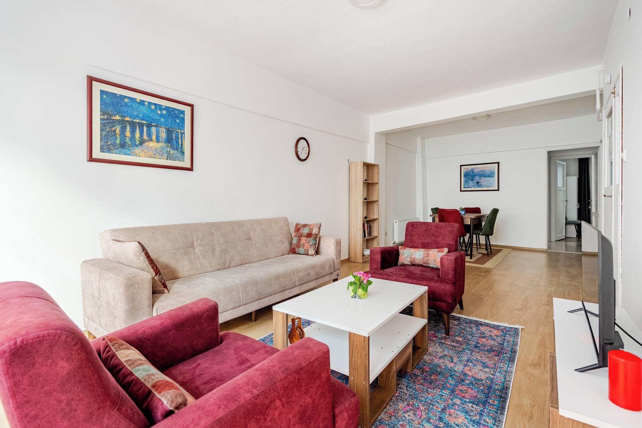 A modern living room with a sofa, armchairs, a coffee table, and a TV. There is a bookshelf and a painting on the wall.