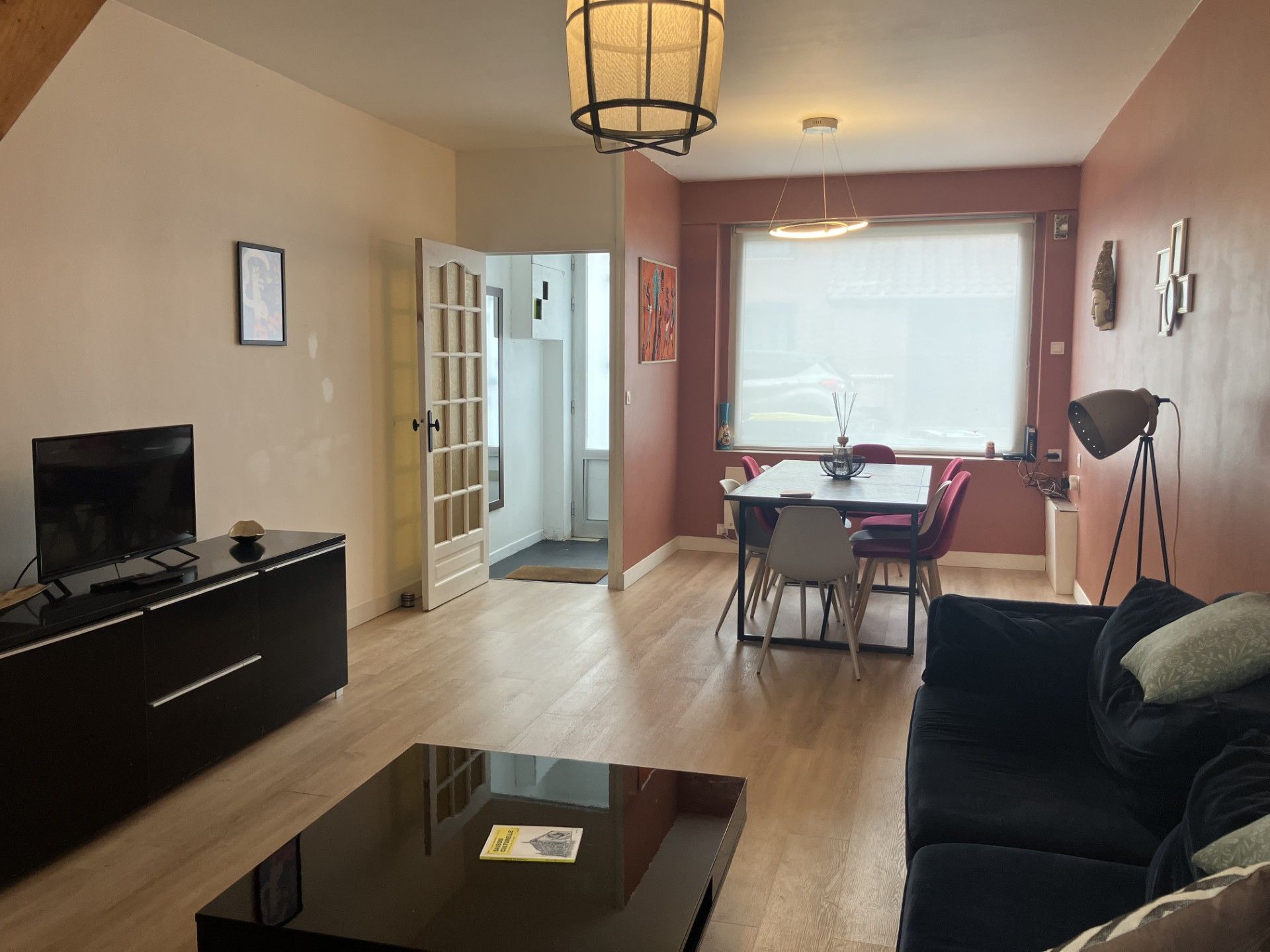 Een woonkamer met een televisie, een zwarte bank en een eethoek met stoelen en een tafel.