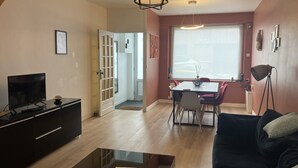 Een woonkamer met een televisie, een zwarte bank en een eethoek met stoelen en een tafel.