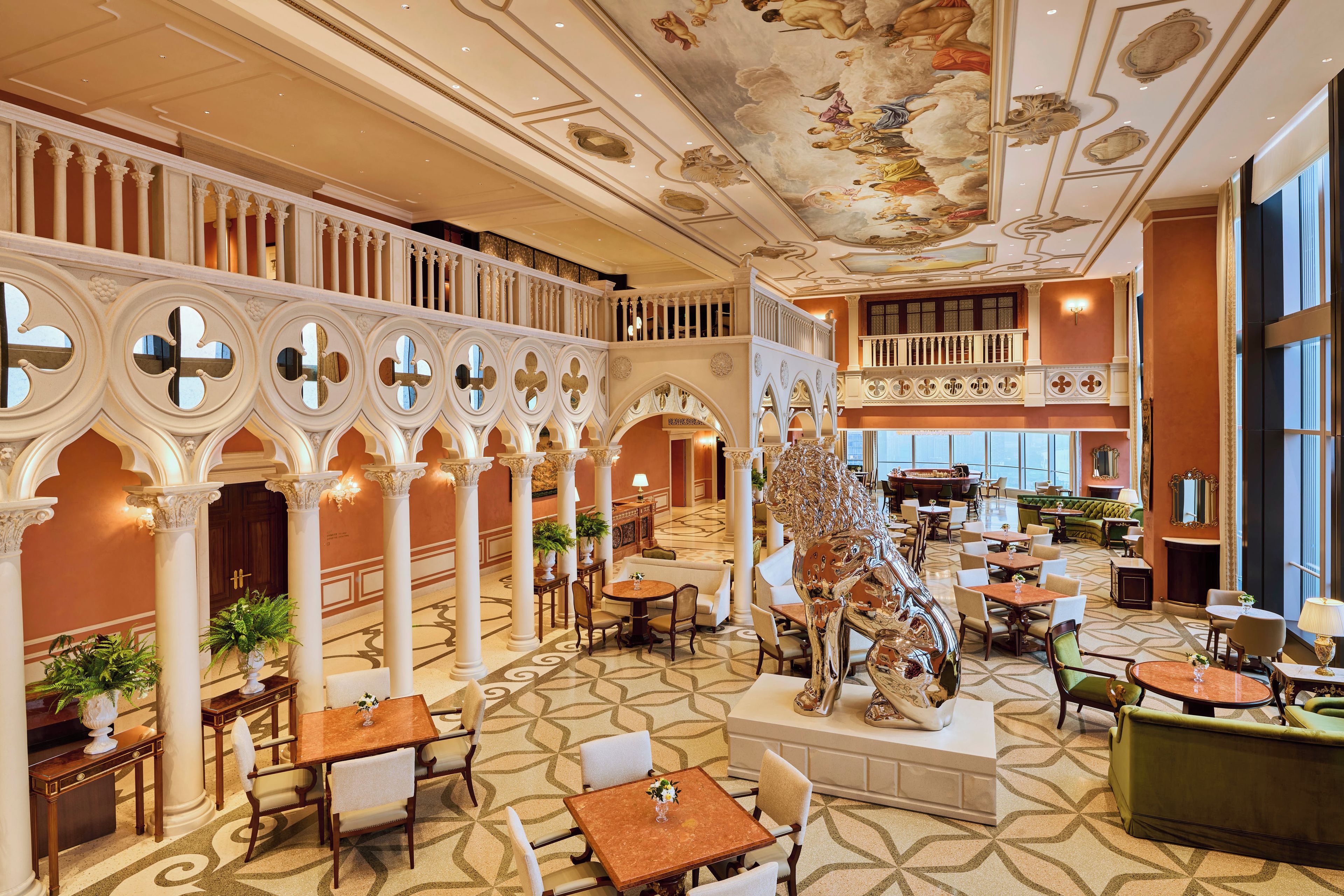 Um hall de hotel espaçoso, com colunas ricamente decoradas, uma escultura central e diversas áreas de estar.