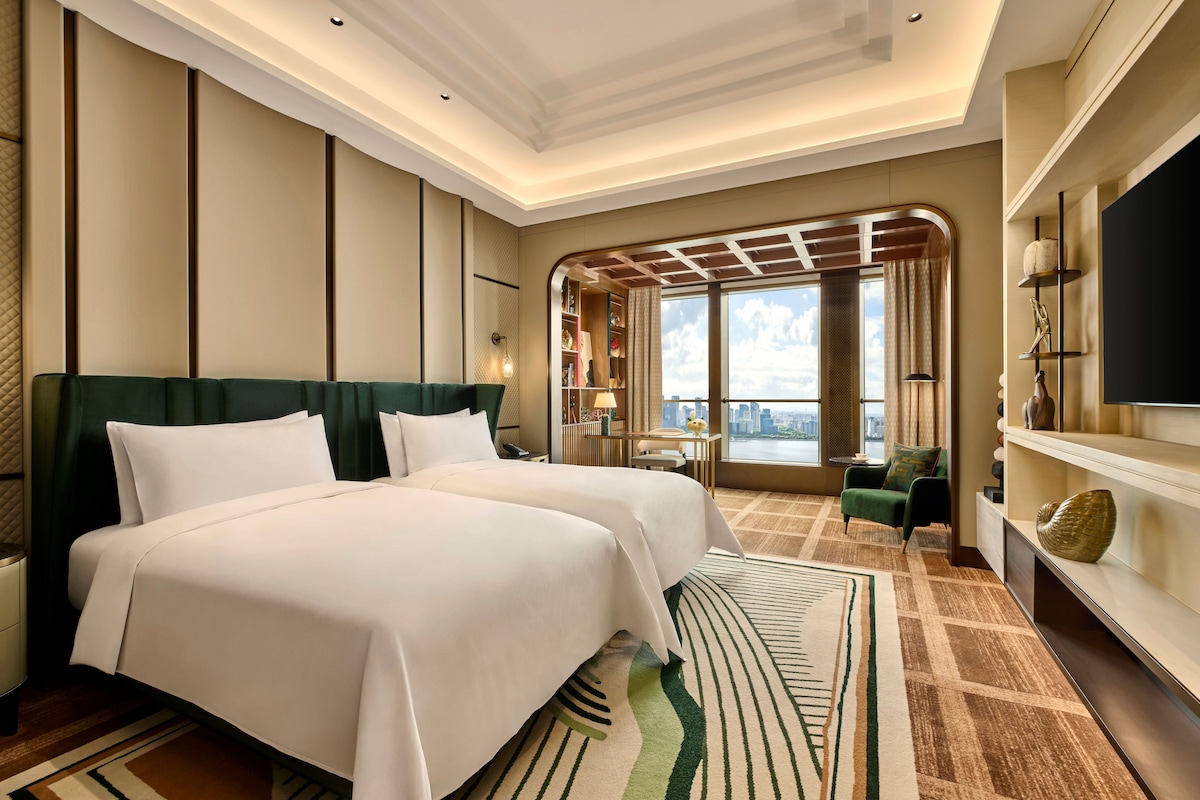 Habitación de hotel con dos camas, un ventanal grande con vista a la ciudad, televisión y un área de descanso con vistas.