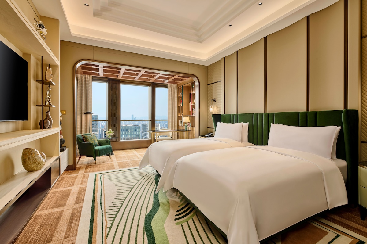 Habitación de hotel con dos camas, un ventanal con vistas, un televisor de pantalla plana y un sillón verde.