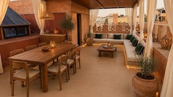 A covered outdoor seating area with a wooden table and chairs, a sofa with cushions, and potted plants.