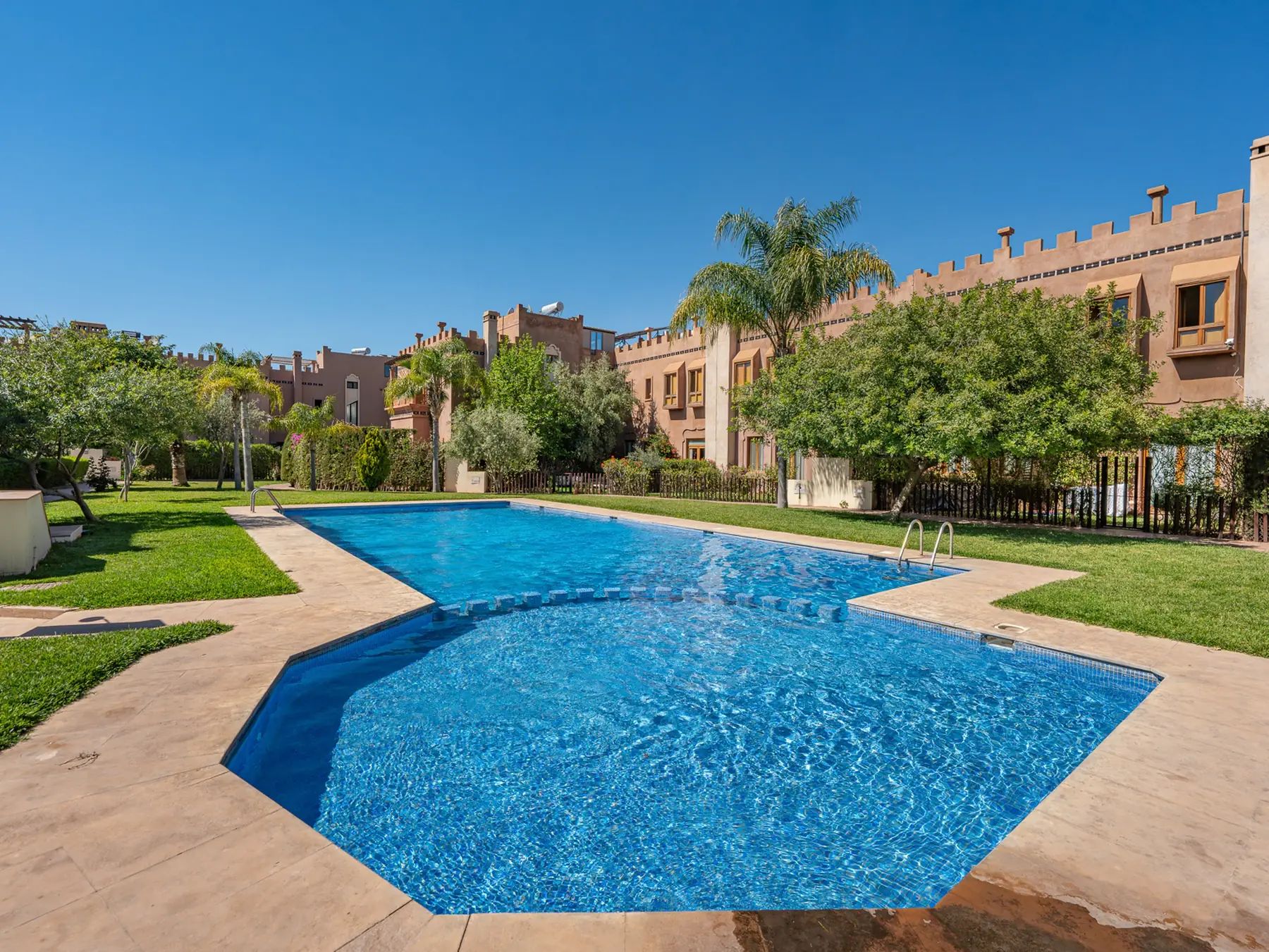 A rectangular swimming pool with a clear blue water surface, surrounded by a paved walkway, and adjacent to a building with a castle-like architectural style.