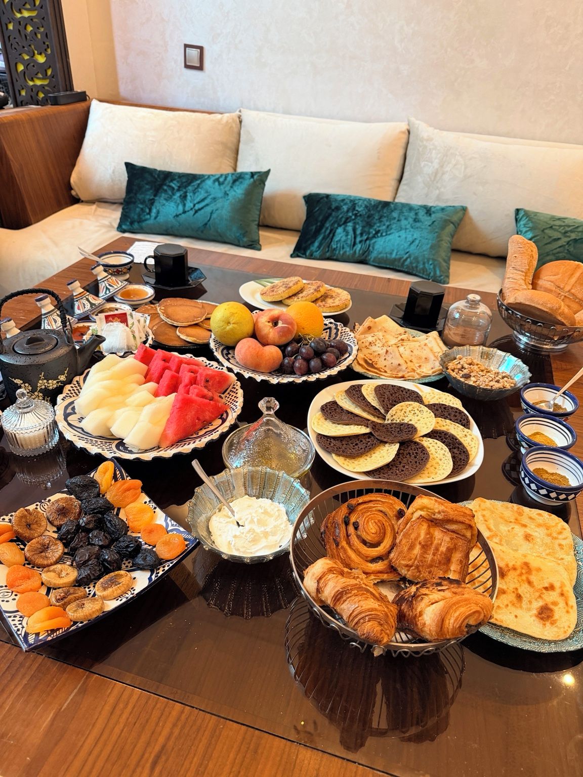 A breakfast buffet with various pastries, fruits, and beverages on a table in a living room with a sofa and cushions.