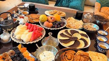A breakfast buffet with various pastries, fruits, and beverages on a table in a living room with a sofa and cushions.