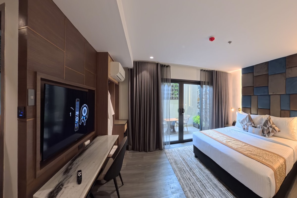 Et moderne hotelværelse med et fladskærms-tv, et skrivebord, en seng med sengegavl og en skydedør til balkonen.