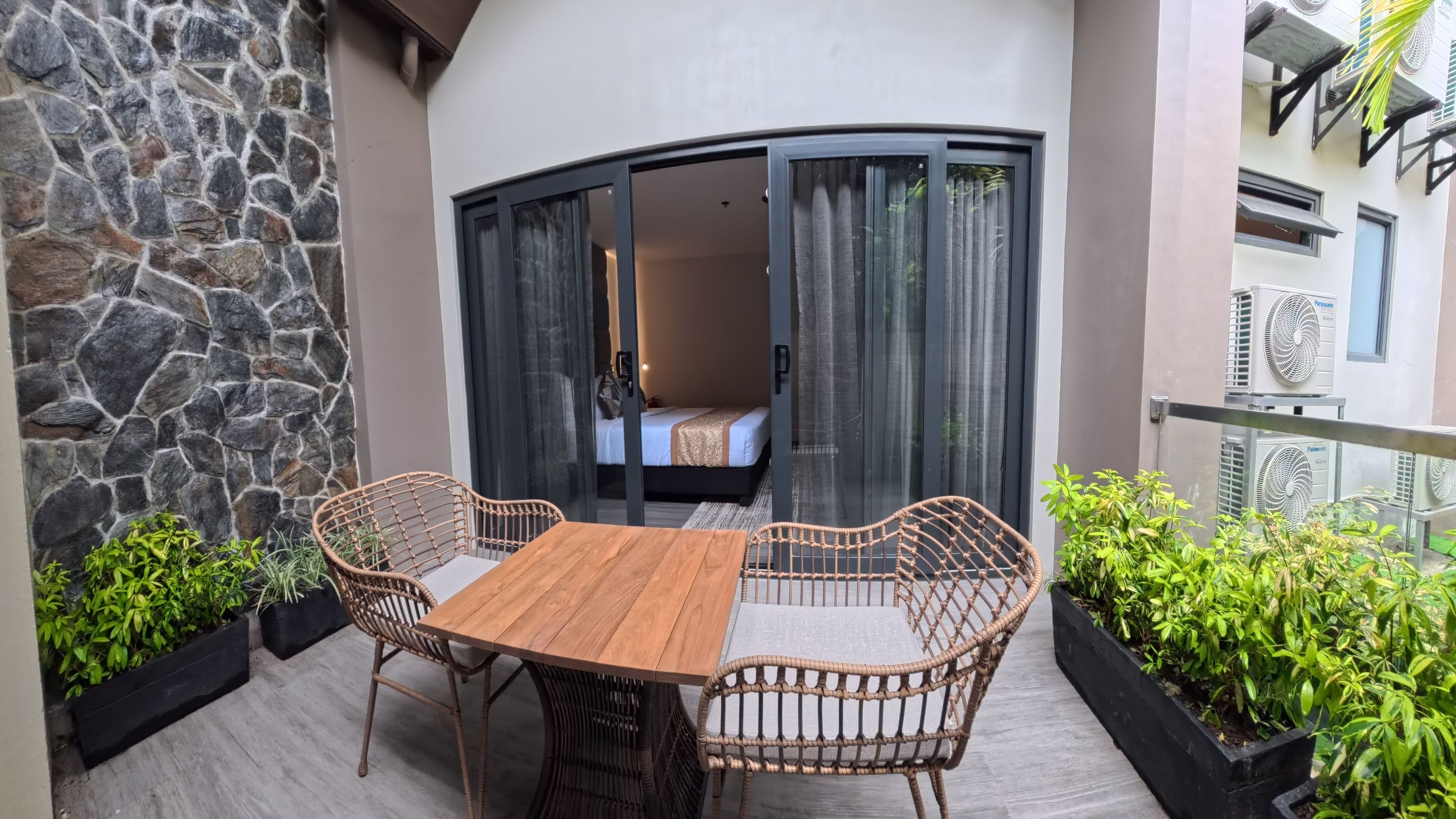 Un balcon avec une table en bois et des chaises en osier, un mur en pierre et une vue sur une pièce avec un lit et des rideaux.