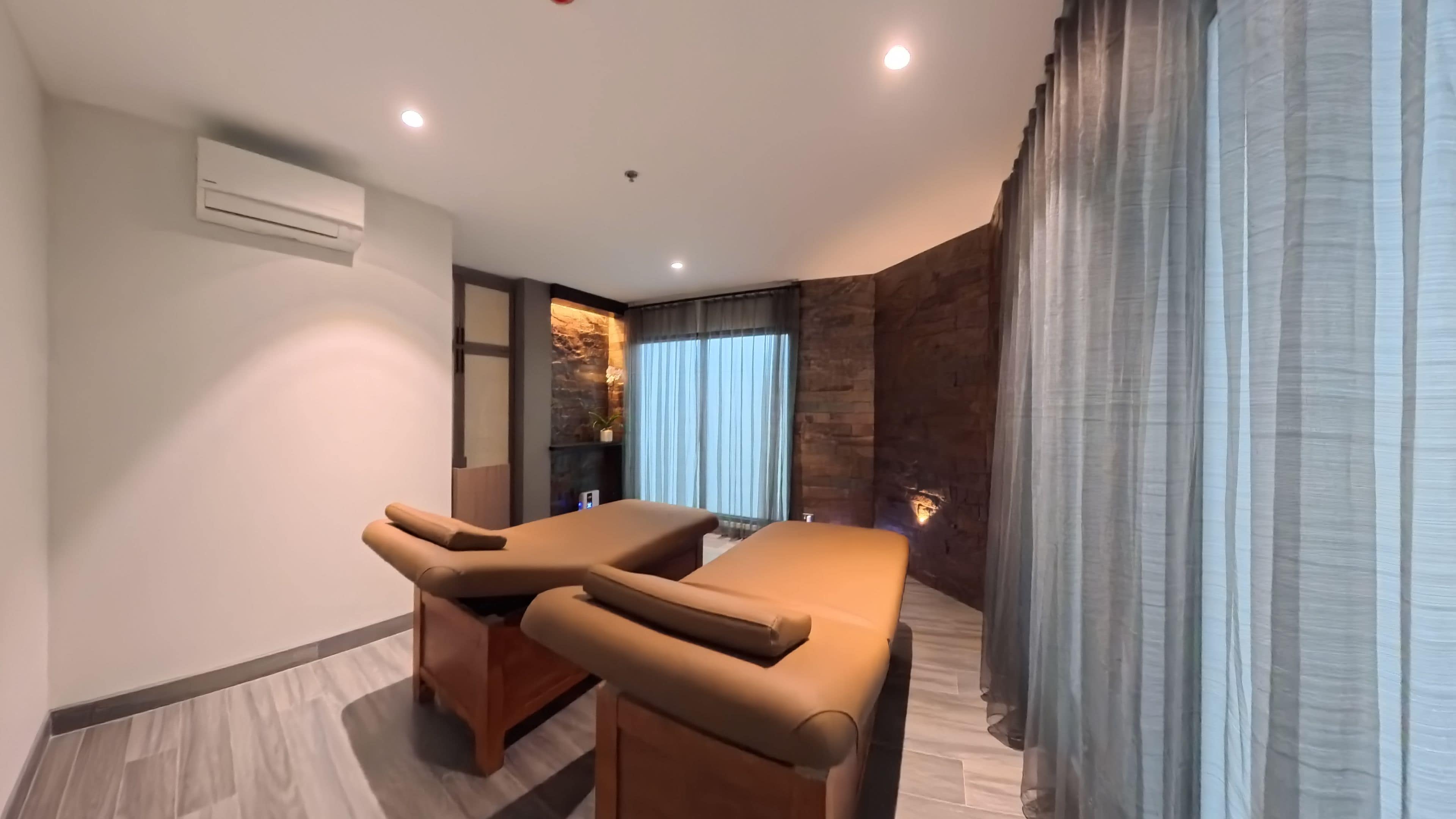 Una habitación con dos camillas de masaje, una pared de acento de piedra y un amplio ventanal con cortinas.