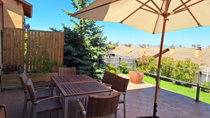 An outdoor patio with a wooden table and chairs, a large umbrella, and a view of neighboring buildings.