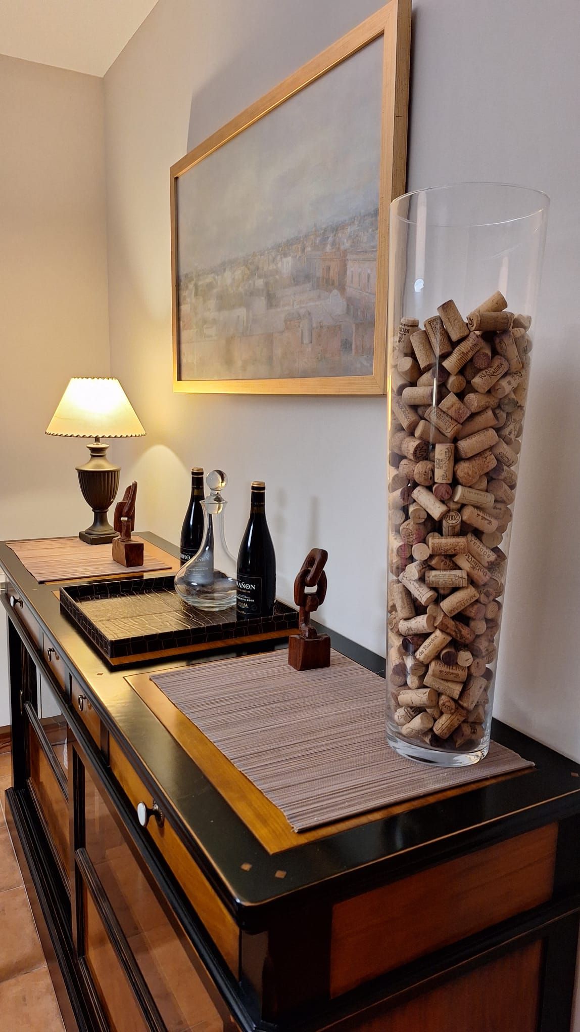 A room with a desk, a lamp, a bottle of wine, a glass bottle, and a vase filled with corks.