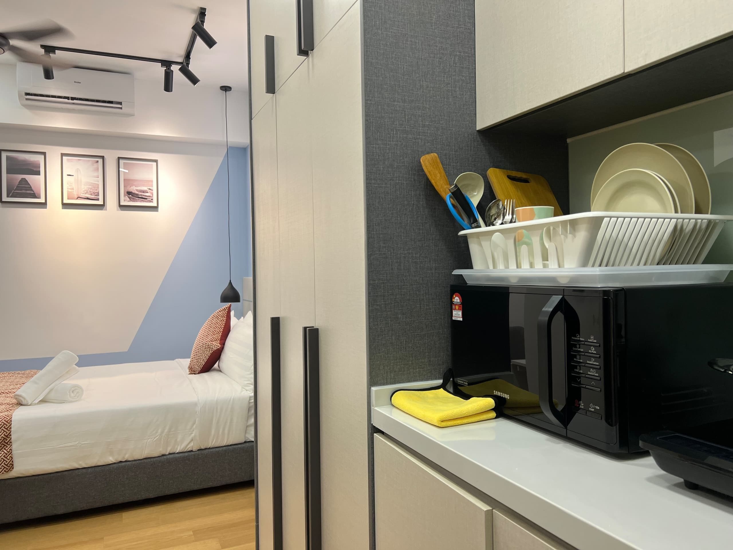 Habitación de hotel compacta con cama, microondas y pequeña cocina equipada con pileta y utensilios.