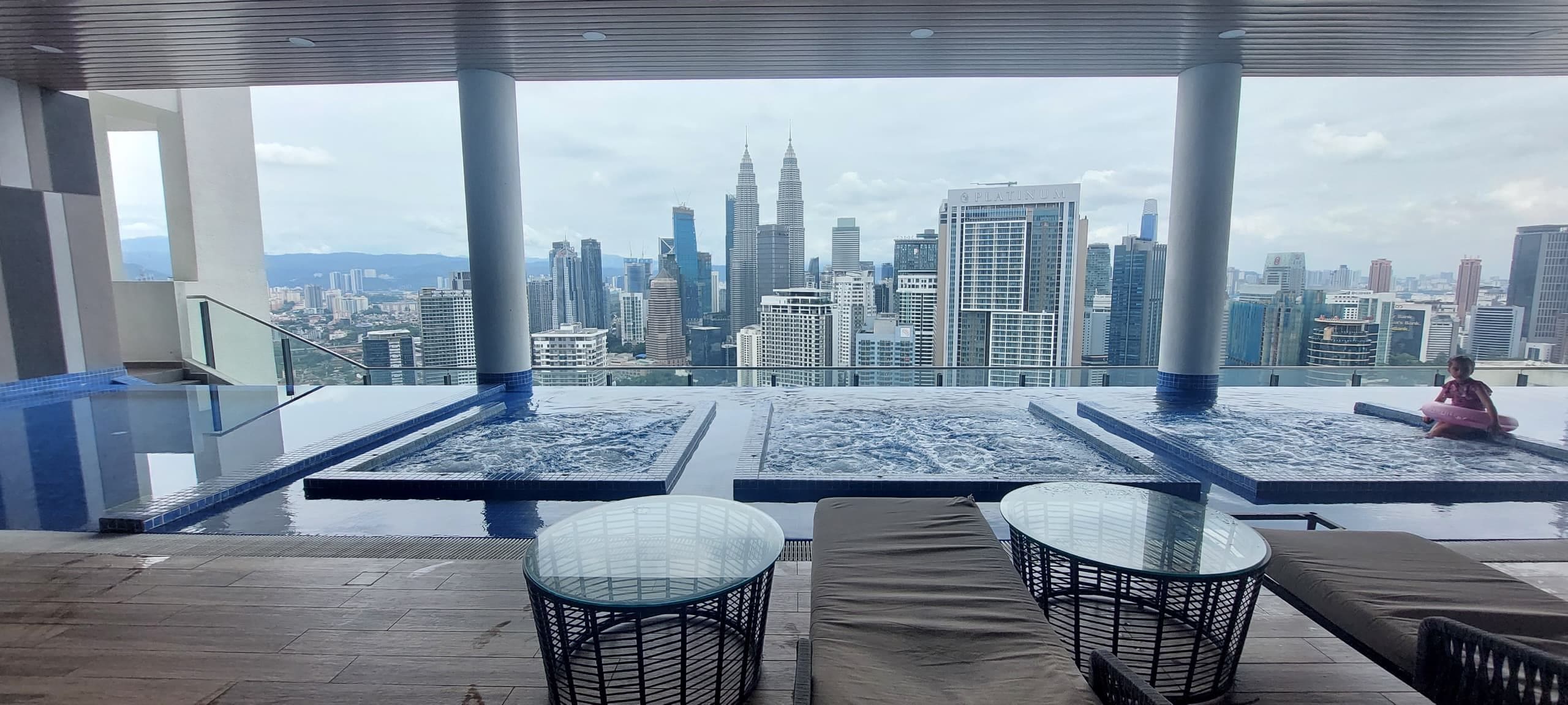 Una piscina en la azotea con vistas a la ciudad, incluyendo las Torres Petronas, y una zona de estar cubierta con vidrio que tiene una mesa.