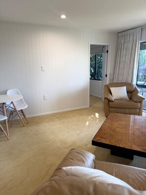 A room with a beige carpet, a brown leather sofa, a white dining table with chairs, and a window with curtains. - Your Stay with a View - a little hidden sanctuary (Taupō)