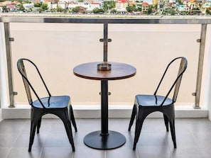 Un balcón con una mesa redonda y dos sillas, barandilla metálica y vista a un paisaje urbano.