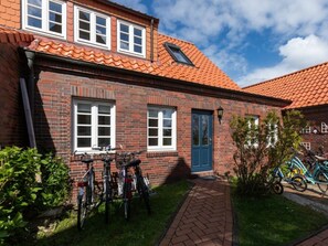 A brick building with a red-tiled roof, a blue door, and a brick pathway leading to a bicycle parking area. - 272 - Apartment Lighthouse (Juist)
