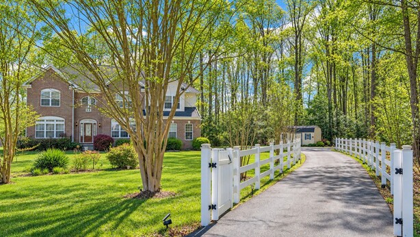 A residential property with a paved driveway, white picket fence, and a brick house with multiple windows. - Private 5BR Estate with Hot Tub, Pool & Movie Room (Hughesville)