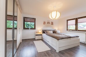 Ein Schlafzimmer mit einem Bett, Nachttischen, einer Lampe, einem Fenster mit Jalousien und einer Deckenleuchte.