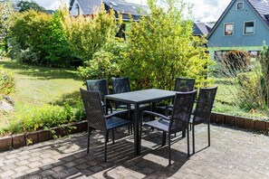 Eine Terrasse im Freien mit Tisch und Stühlen, umgeben von Grünpflanzen und einem Haus im Hintergrund.