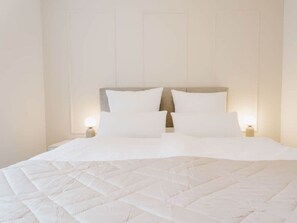 Una cama con edredón blanco acolchado y dos almohadas, una mesita de noche con lámpara, y un panel de pared decorativo.