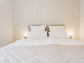 Una cama con edredón blanco acolchado y dos almohadas, una mesita de noche con lámpara, y un panel de pared decorativo.