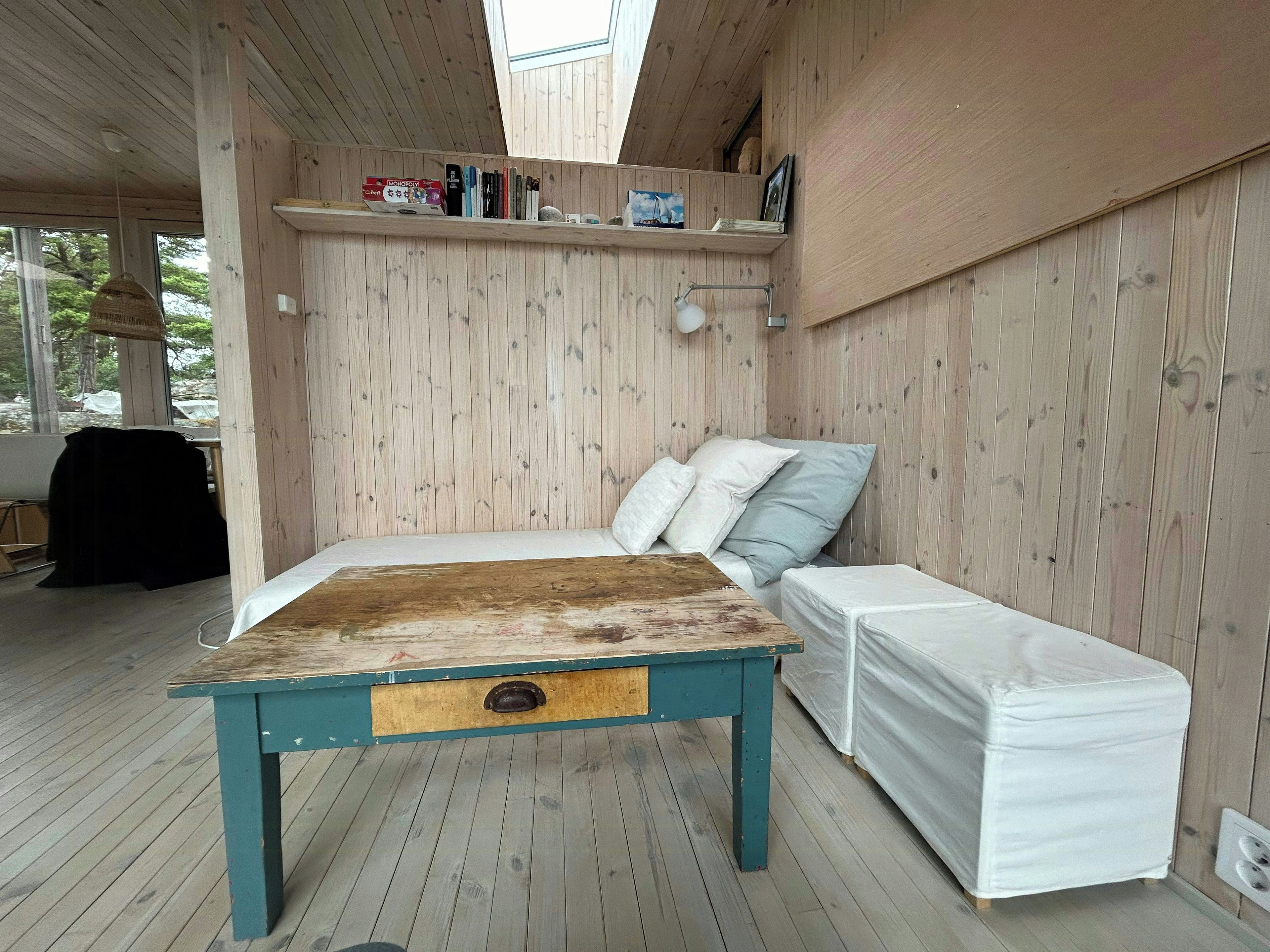 Ein Zimmer aus Holz mit einem Bett, einer Bank, einem Tisch und einem Regal mit Büchern und einem Gesellschaftsspiel.