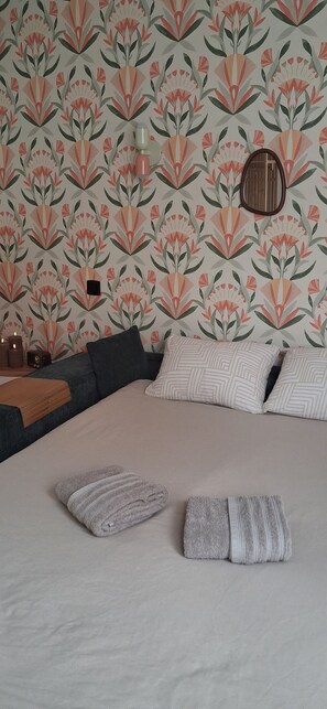 Ein Schlafzimmer mit ornamentalem Wandpapier, einem Bett mit Kissen, einem Nachttisch mit Lampe und einem an der Wand befestigten Spiegel.