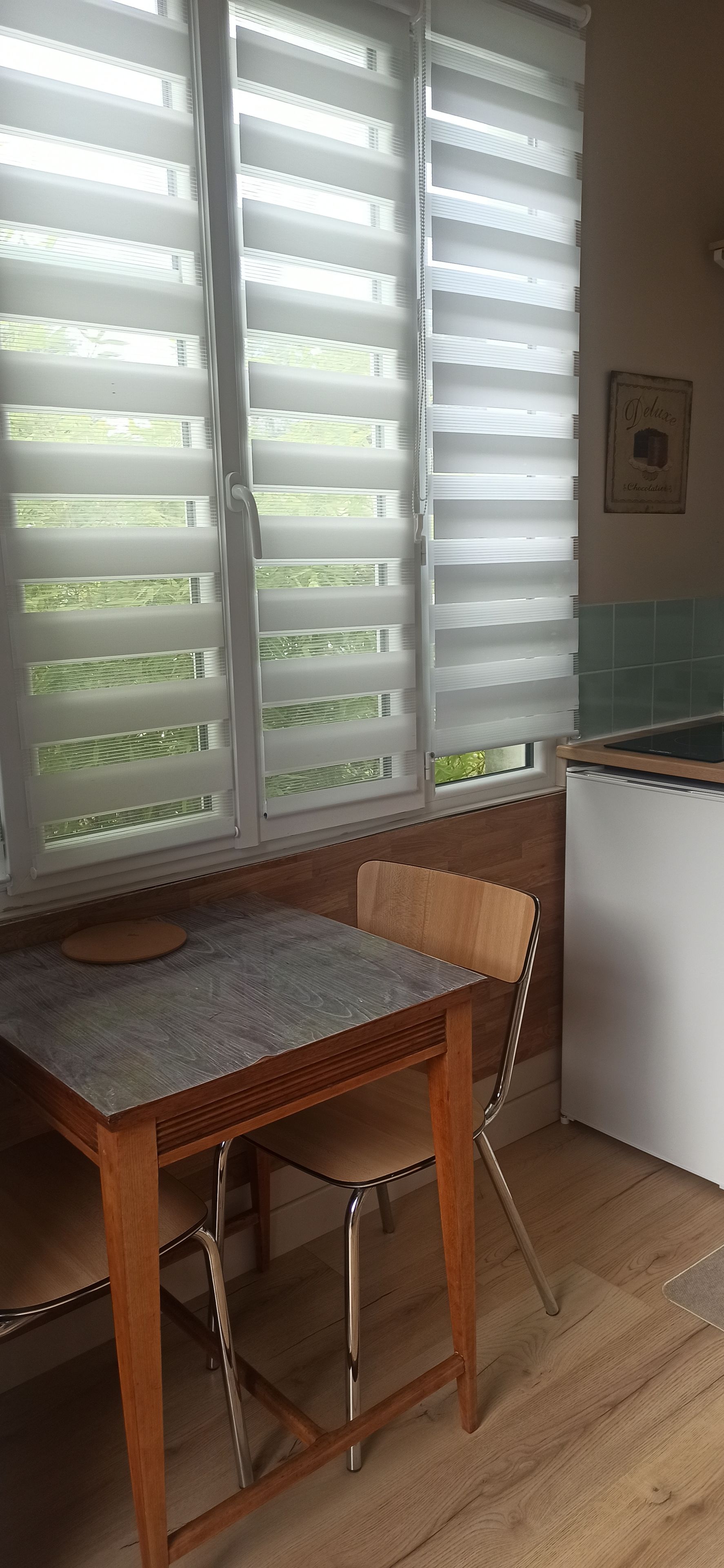 Une cuisine équipée d’une table et de chaises, d’un réfrigérateur et d’une fenêtre avec des stores.