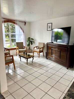 Ein Wohnzimmer mit Fliesenboden, Holzmöbeln, einem Fernseher und einem Fenster mit Vorhängen.