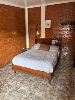 A bedroom with a brick wall, a wooden bed frame, a nightstand with a lamp, and a framed picture on the wall.