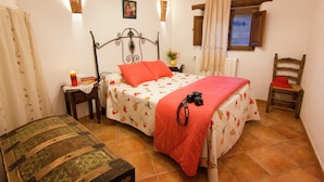 Quarto com cama, mesa de cabeceira, cadeira, baú de madeira e janela com cortinas.