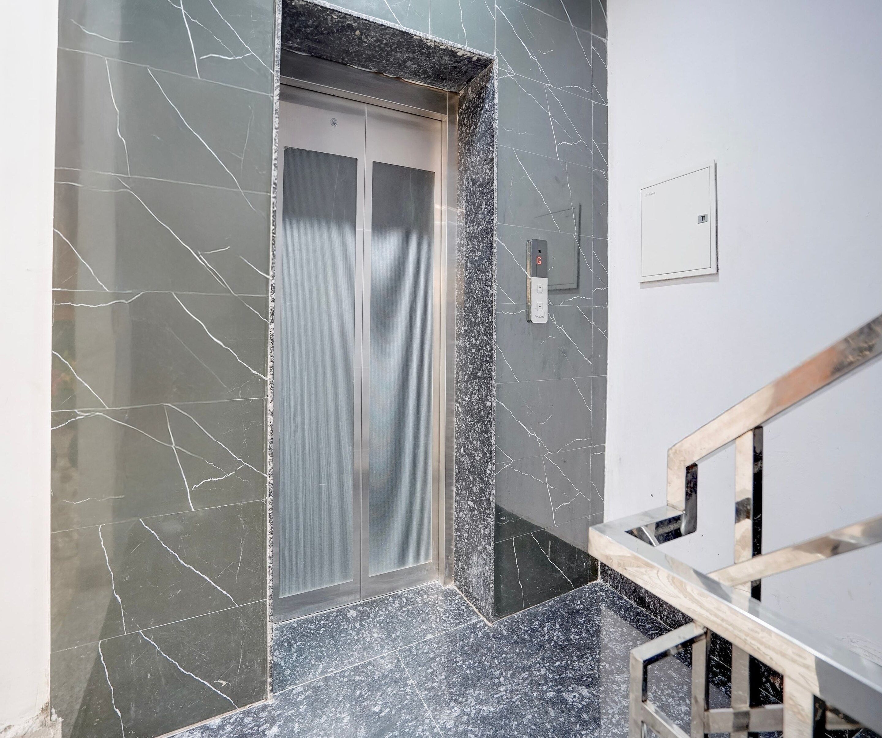 Un ascenseur doté d’une porte en verre, un mur de marbre aux veines blanches et un escalier en bois avec une rampe métallique.