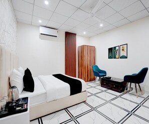 A hotel room with a bed, a desk with a coffee machine, a wardrobe, a blue chair, and a wall with framed artwork. - Classic Basai Chowk (Gurugram)