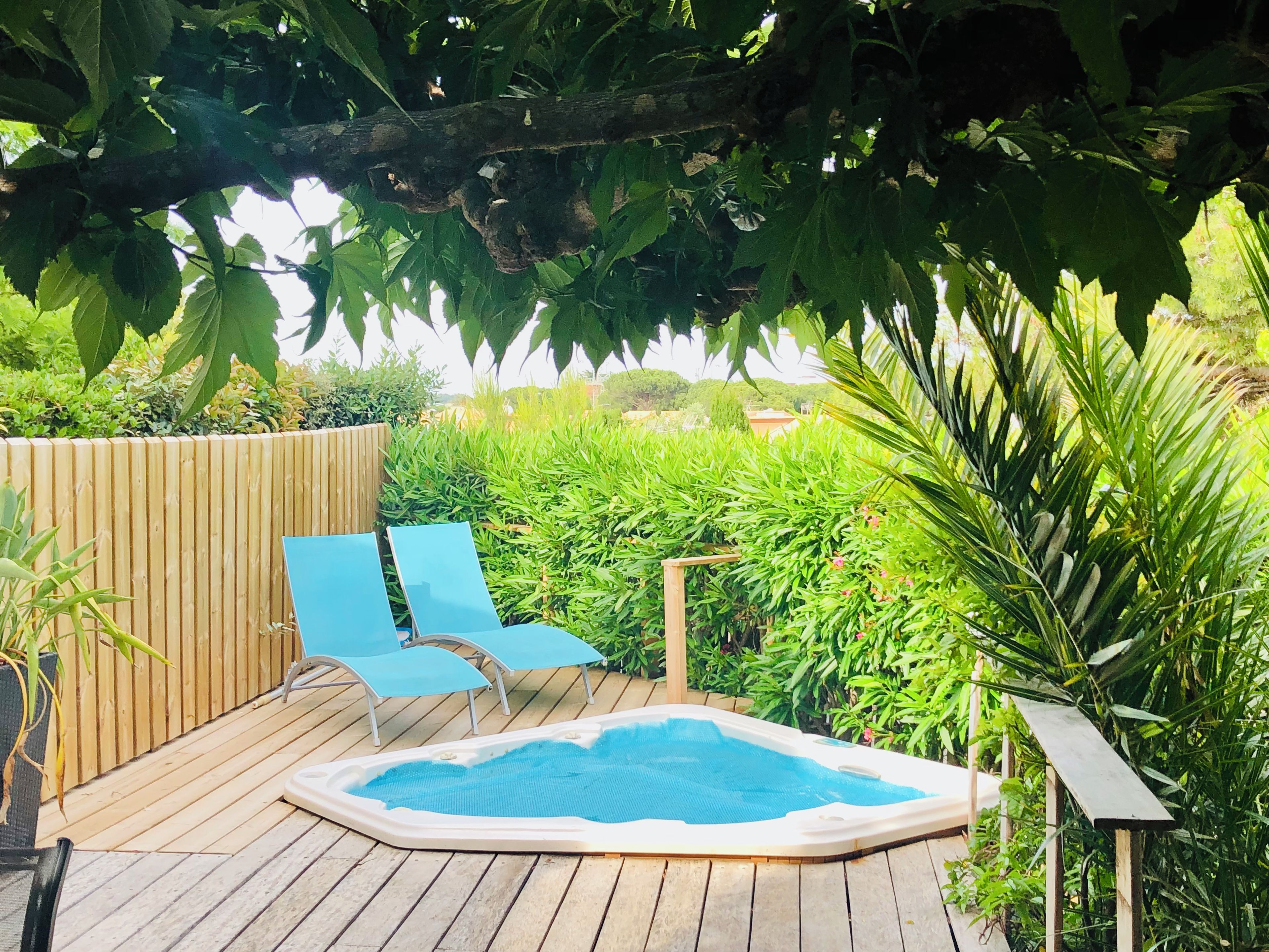 A wooden deck with a hot tub, blue lounge chairs, and lush greenery.