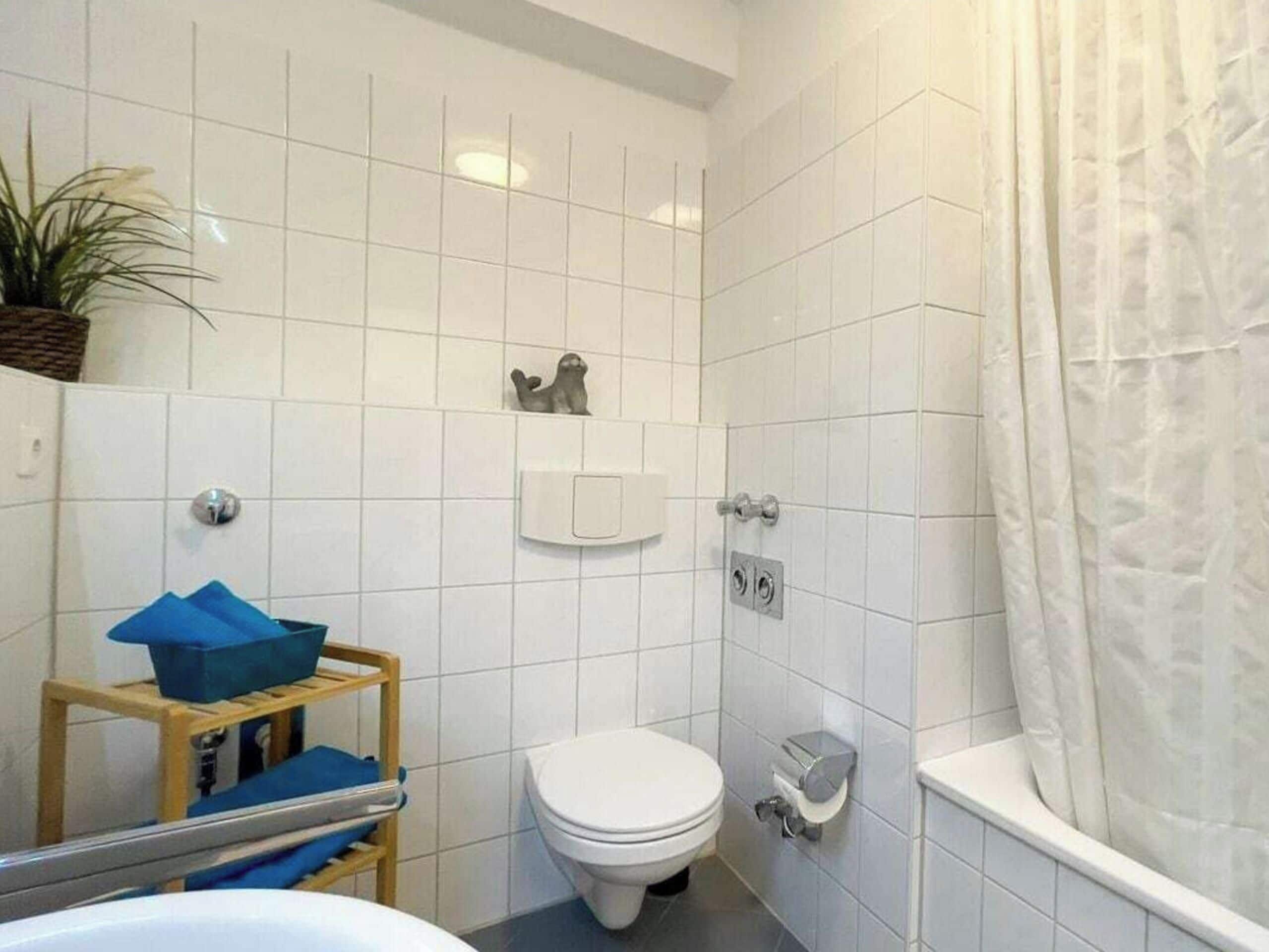 A bathroom with white tiles, a toilet, a shower area, and a small shelf with a blue object.