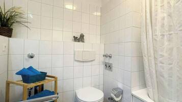 A bathroom with white tiles, a toilet, a shower area, and a small shelf with a blue object.