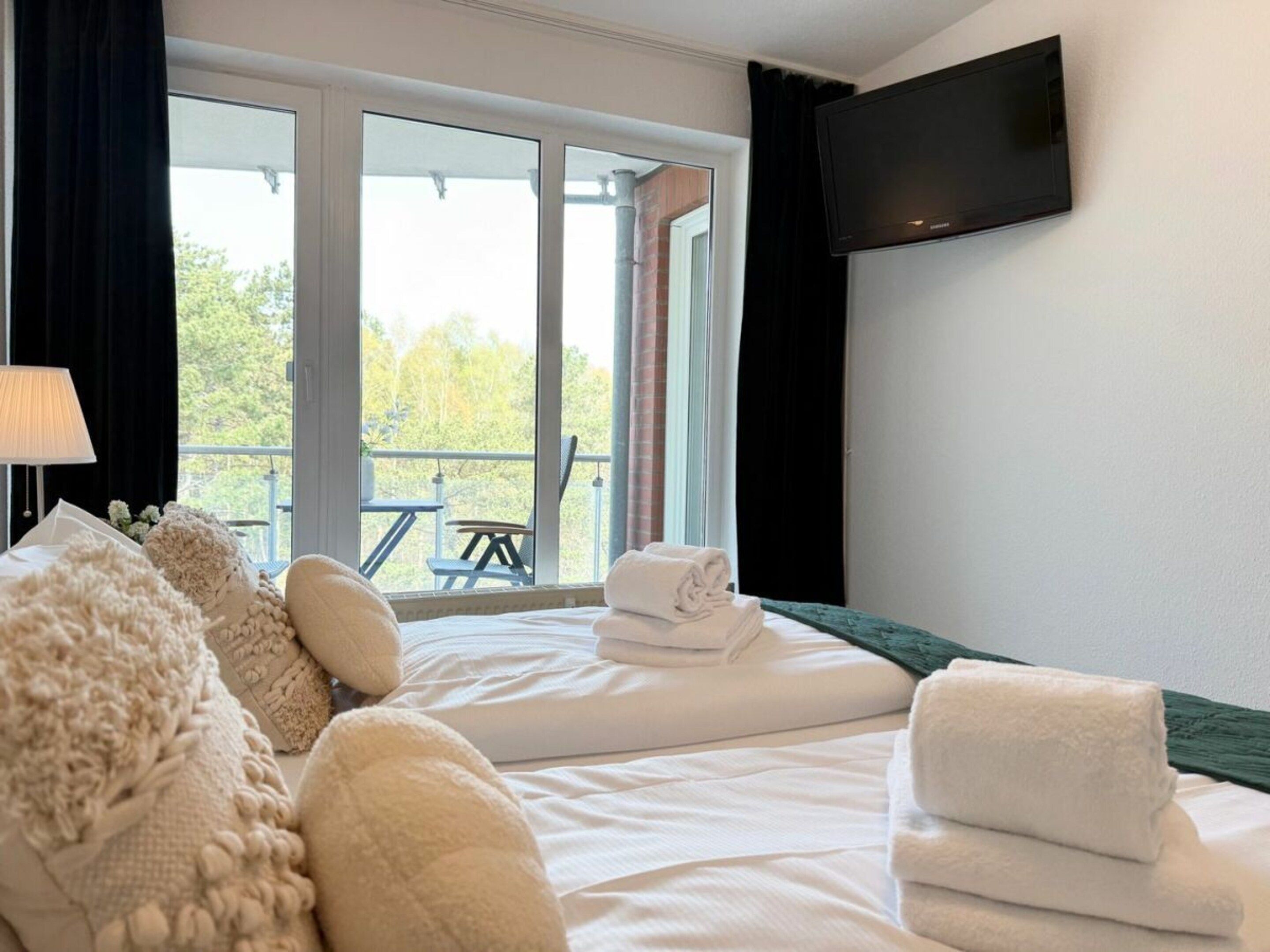 A hotel room with a bed, a TV mounted on the wall, and a balcony with outdoor seating.