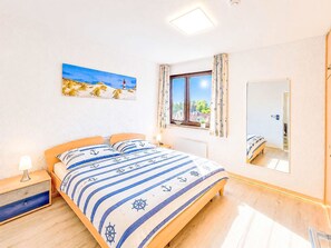 Ein Schlafzimmer mit einem Bett, einem Nachttisch, einem Fenster mit Vorhängen, einem Bild von einer Strandlandschaft und einem Spiegel.