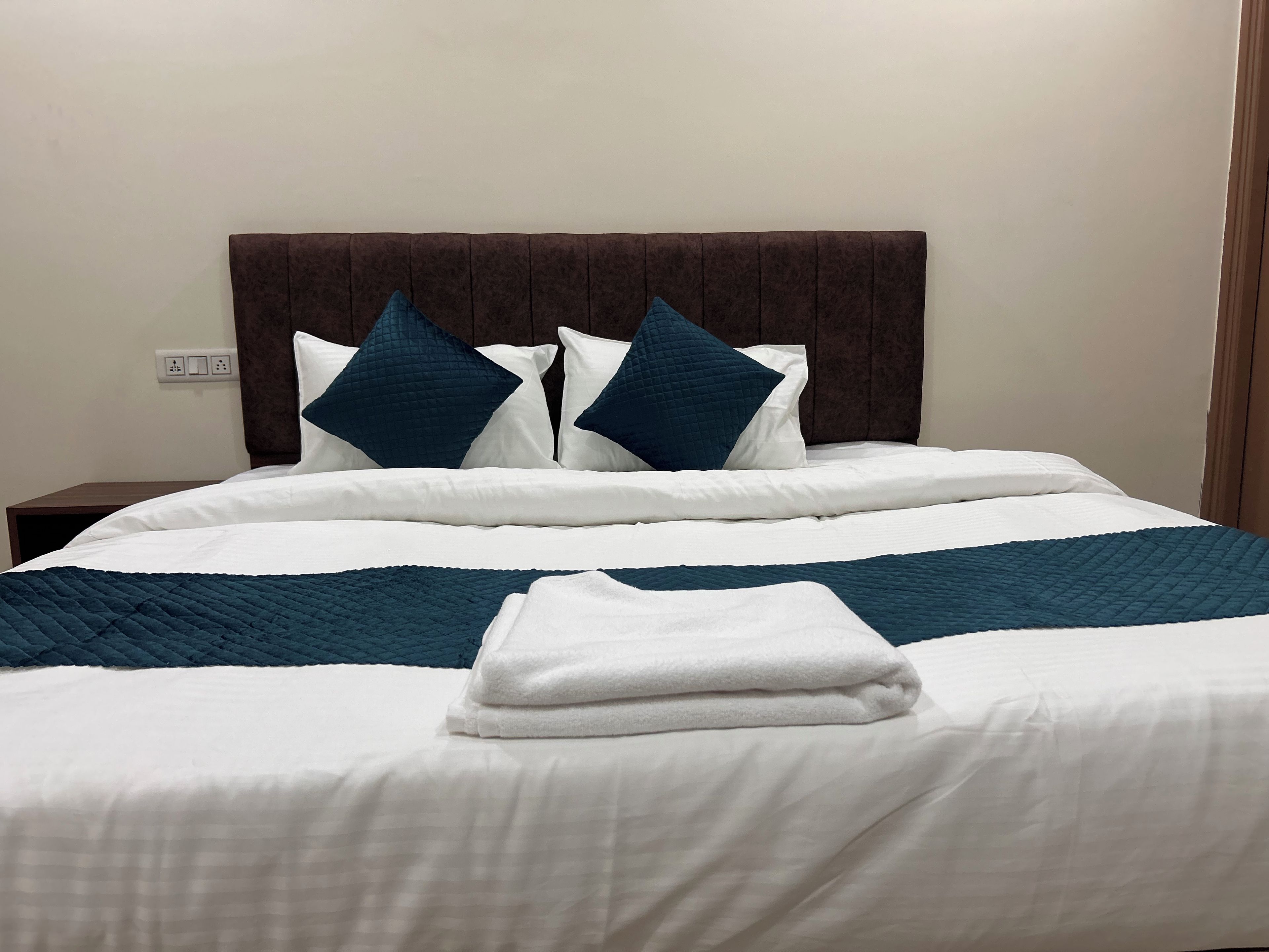 Een net opgemaakt bed met een donker hoofdbord, witte lakens en een gevouwen witte handdoek.